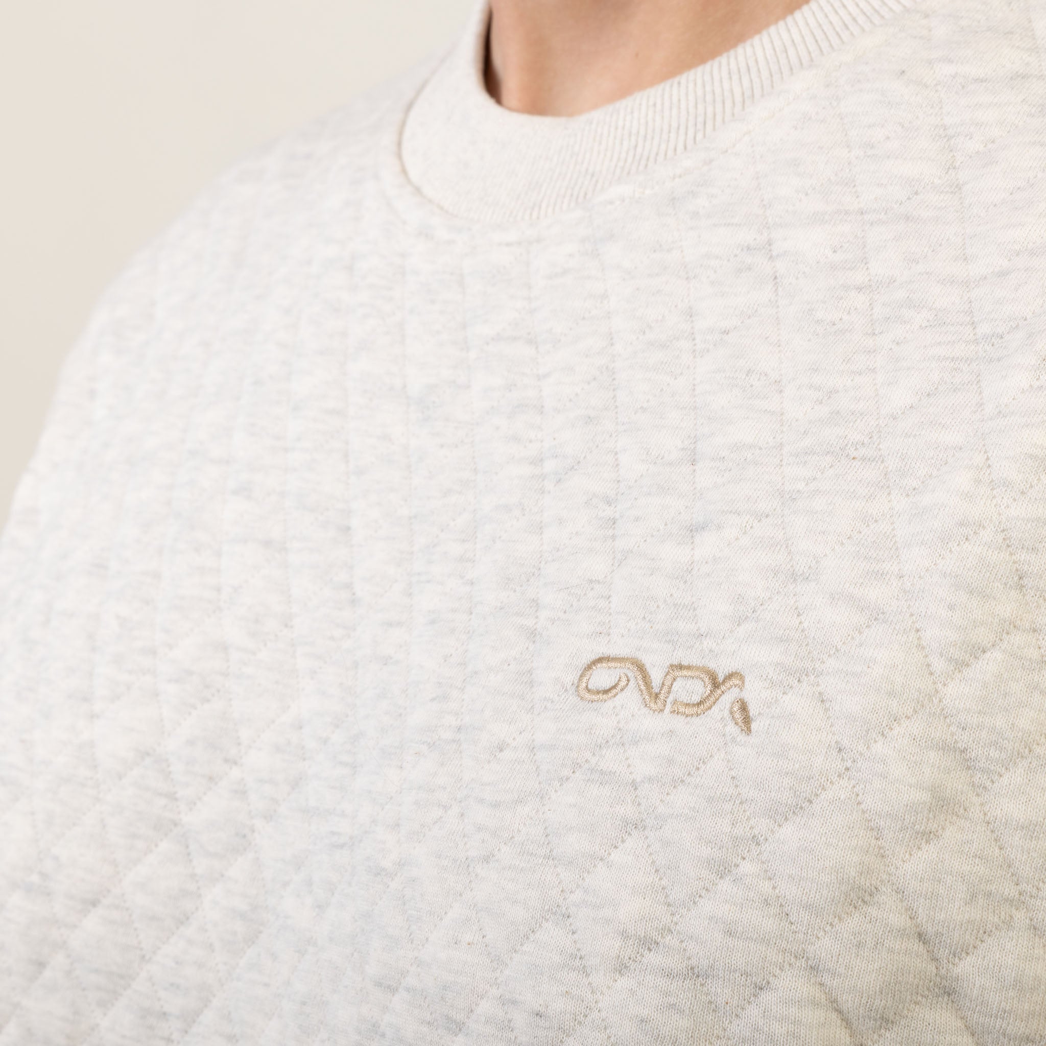 Close-up of a white sweater with a gold embroidered onda logo on a beige background